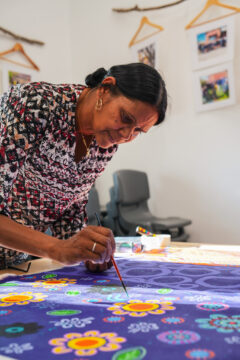 A woman leans over a table while painting colorful floral designs on a purple canvas. She is focused, holding a paintbrush, with framed photos and hangers on the wall behind her.