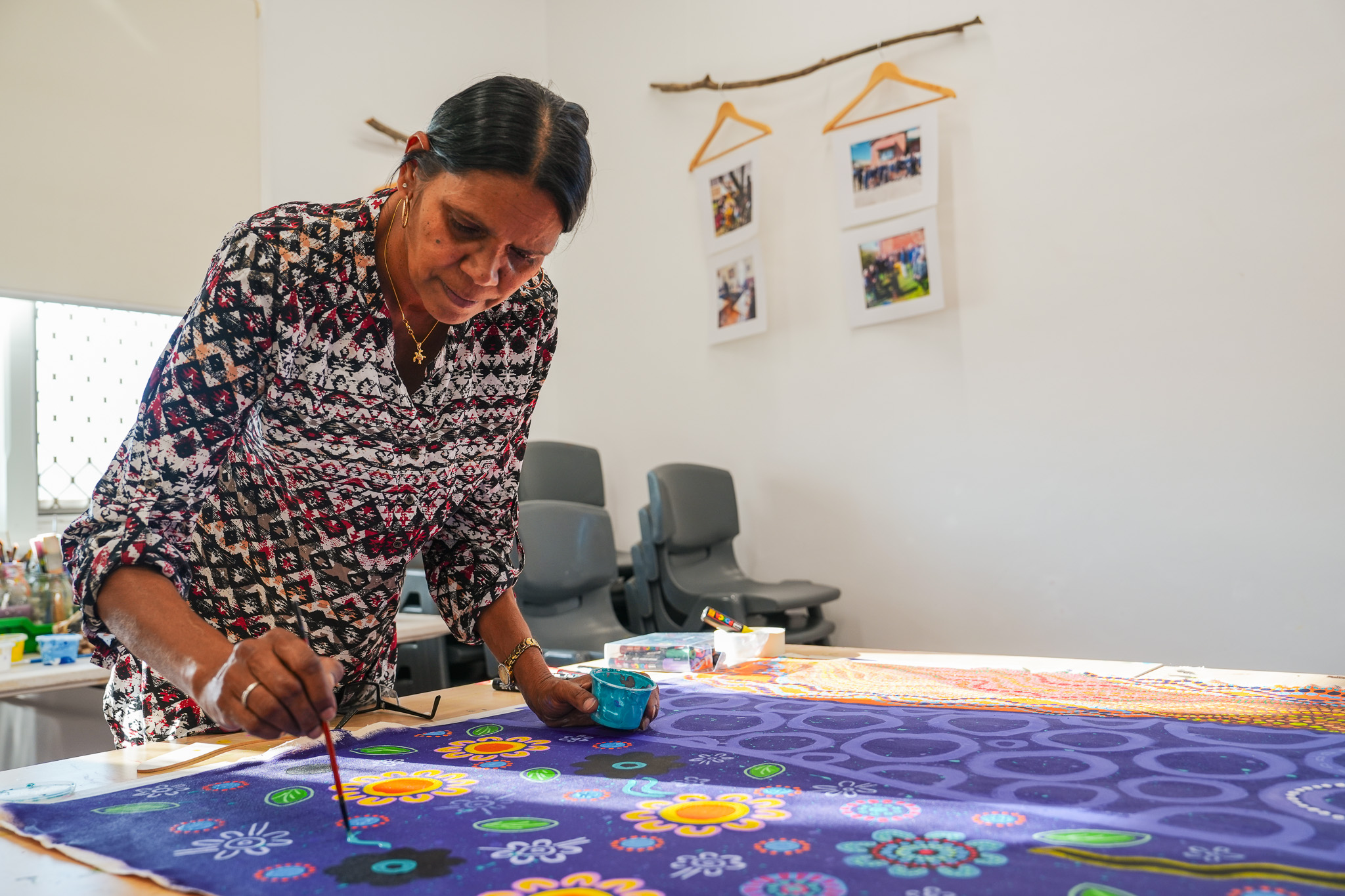 A woman paints colorful floral patterns on a large piece of fabric in a bright, modern room with framed photos and empty chairs in the background.