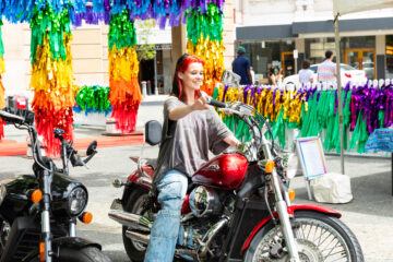 A person with red hair and ripped jeans smiles while sitting on a red motorcycle at an outdoor event, with colorful rainbow streamers hanging in the background.