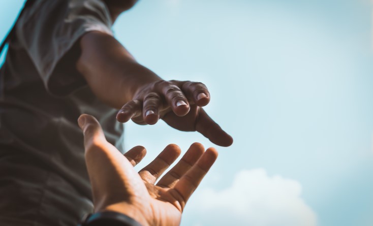 A close-up of two hands reaching toward each other against a bright sky, with one hand reaching down and the other reaching up, symbolizing help, support, or connection.