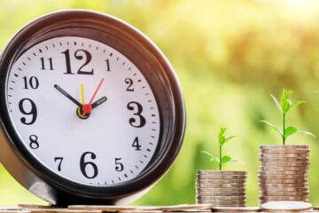 A clock showing 12:00 sits next to two stacks of coins, each topped with a small green plant, symbolizing growth over time. The background is blurred with natural green foliage.