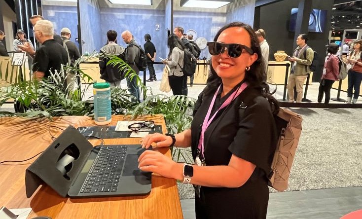 A woman wearing sunglasses and a backpack smiles while using a keyboard and tablet at a wooden table in a busy indoor event or conference setting. People are gathered and walking in the background.