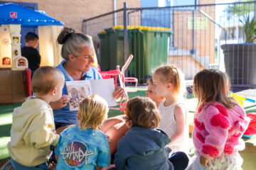 A woman sits outside showing a picture book to a group of young children, who are gathered around her and listening attentively. They are in a sunny playground area with toys and a fence visible in the background.