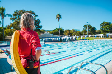A lifeguard in a red shirt stands poolside with a yellow floatation device, watching over a large outdoor swimming pool with lane dividers on a sunny day, surrounded by trees and blue sky.