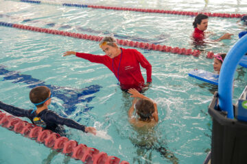 A swim instructor in a red shirt stands in a pool, guiding two children as they practice swimming. Other swimmers and an instructor are visible in nearby lanes, and the pool is divided by lane markers.