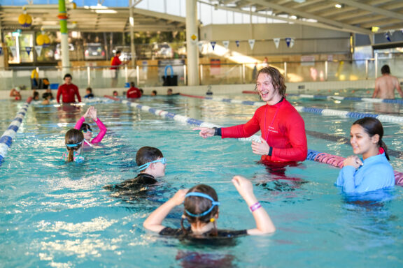 A swim instructor in a red shirt smiles and gestures while teaching a group of children in an indoor swimming pool. The children wear goggles and swimming attire, practicing strokes near the pools lane dividers.