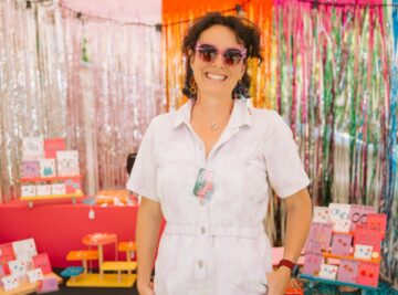 A smiling person in a white outfit and pink sunglasses stands in front of a vibrant, colorful booth with shiny tinsel curtains and displays of art prints and accessories.