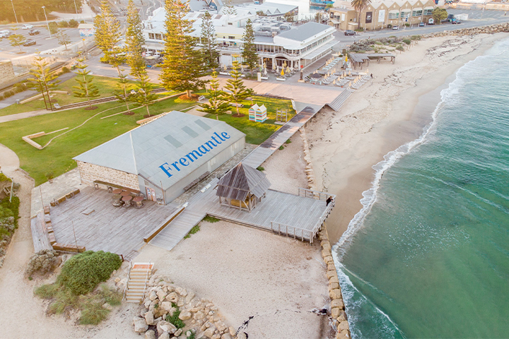 Aerial view of a beachside area in Fremantle, featuring a building with Fremantle written on the roof, wooden boardwalks, green lawns, and gentle waves along a sandy shoreline.