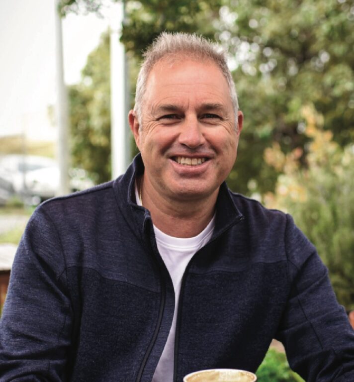 A middle-aged man with short gray hair, wearing a navy jacket and white shirt, smiles while sitting outdoors at a table with a coffee cup. Green trees and blurred cars are visible in the background.