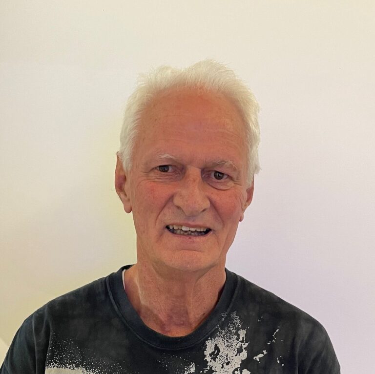 An older man with short white hair is smiling gently at the camera. He is wearing a dark t-shirt with a white splatter pattern and standing in front of a plain, light-colored background.