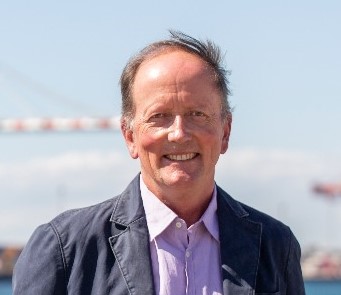 A middle-aged man with short, light brown hair and a receding hairline smiles outdoors. He is wearing a light purple shirt and a dark blazer, with a blurred bridge and blue sky in the background.