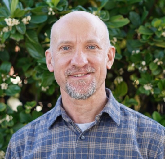 A smiling, bald man with a short beard and blue eyes wearing a plaid shirt stands in front of green foliage with small white flowers.