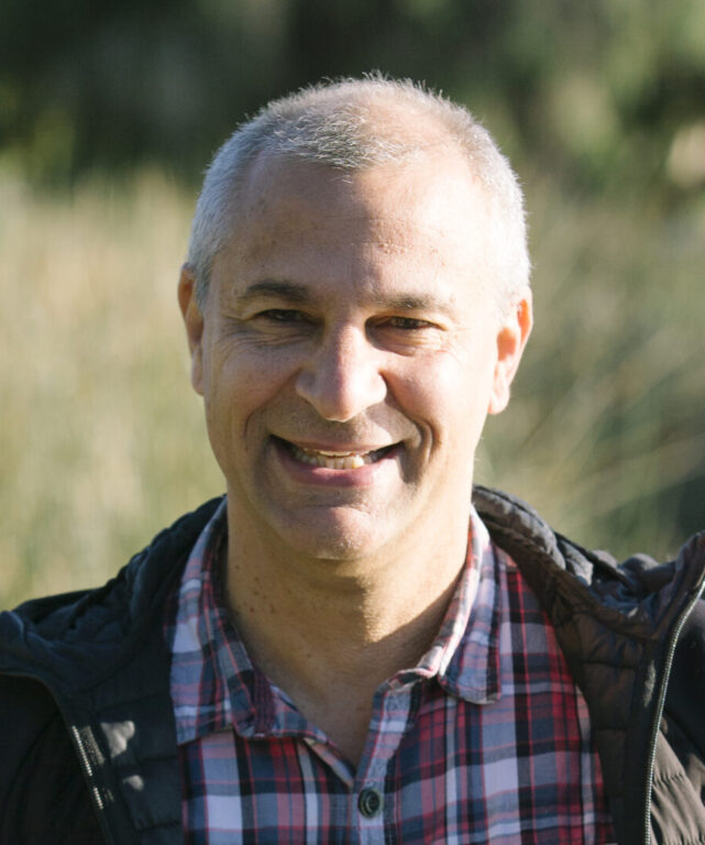 A smiling man with short gray hair stands outdoors, wearing a red plaid shirt and a black jacket. He is leaning on a wooden railing with greenery and blurred trees in the background.