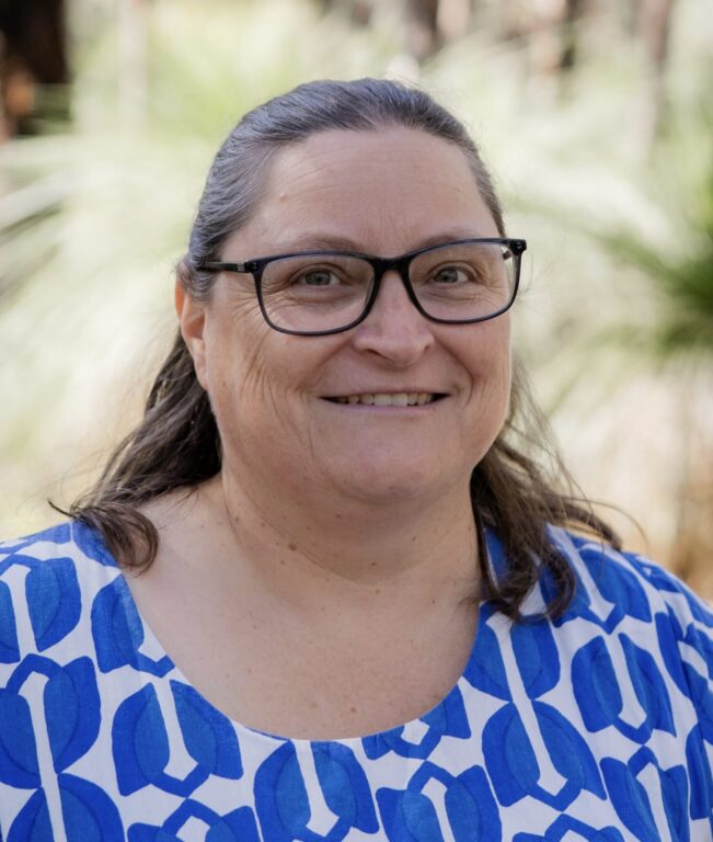 A woman with long brown hair, wearing glasses and a white and blue patterned top, smiles outdoors with greenery in the blurred background.