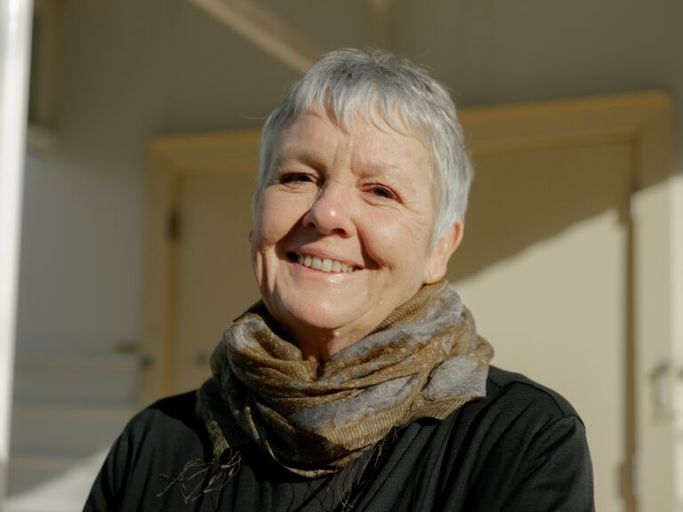 An older woman with short gray hair smiles warmly at the camera. She wears a brown scarf and a black top, standing in front of a beige wall in natural sunlight.