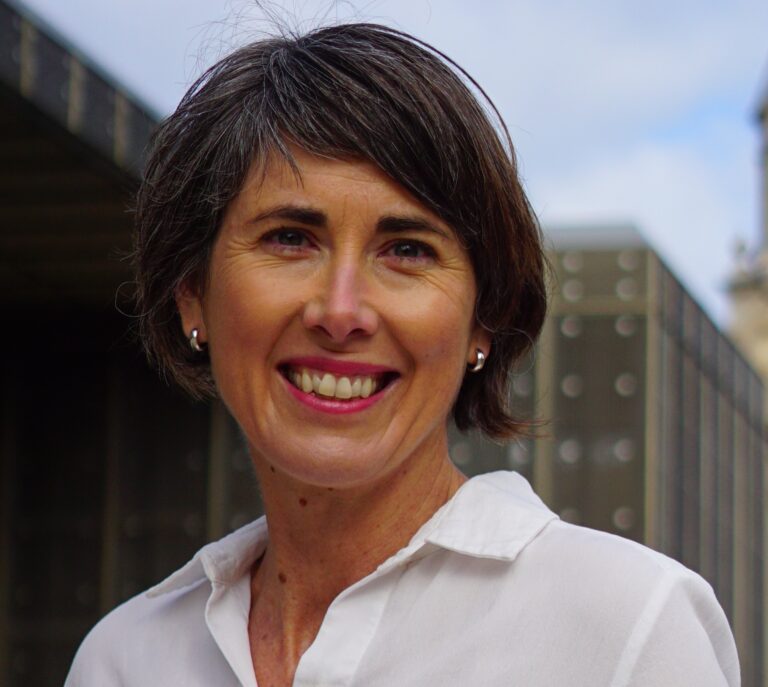 A woman with short gray hair, wearing a white collared shirt and small stud earrings, smiles at the camera outdoors with a blurred modern building in the background.