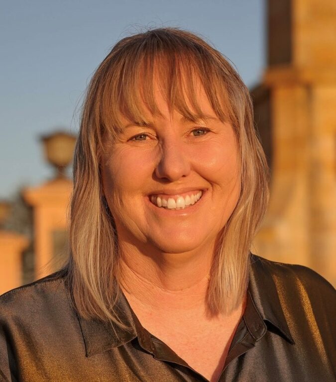 A woman with straight, light-blonde hair and bangs smiles warmly at the camera. She is wearing a short-sleeved, dark, satin-like button-up shirt and is outdoors in golden sunlight, with blurred buildings in the background.