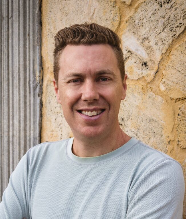 A smiling man with short, light brown hair stands in front of a textured stone wall. He is wearing a light-colored, long-sleeved shirt.