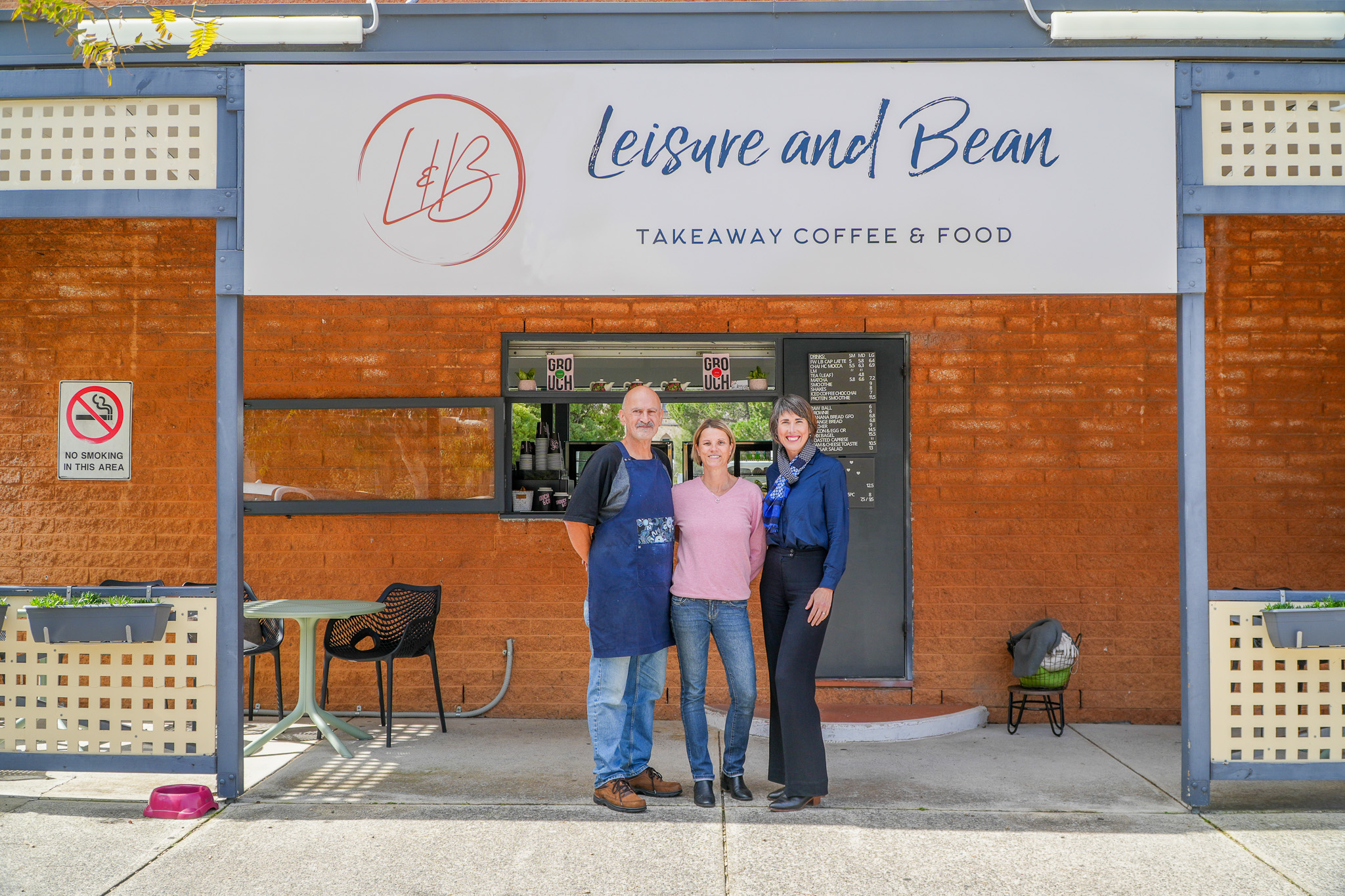 Three people stand smiling in front of a café called Leisure and Bean Takeaway Coffee & Food, with a menu in the window behind them and a No Smoking sign on the left.