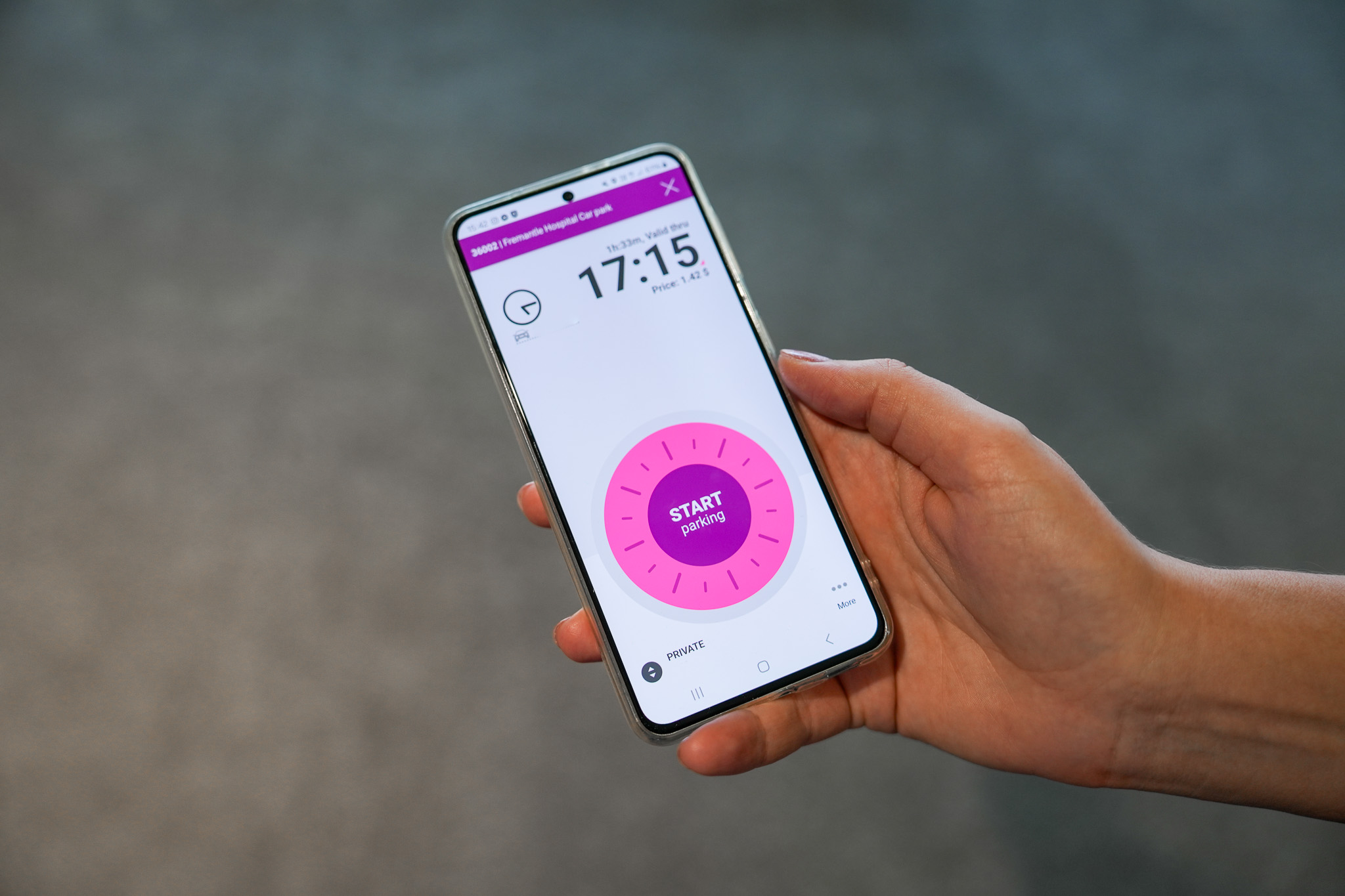A hand holds a smartphone displaying a parking app. The screen shows the current time, parking duration, cost, and a large pink button labeled START parking. The background is blurred.