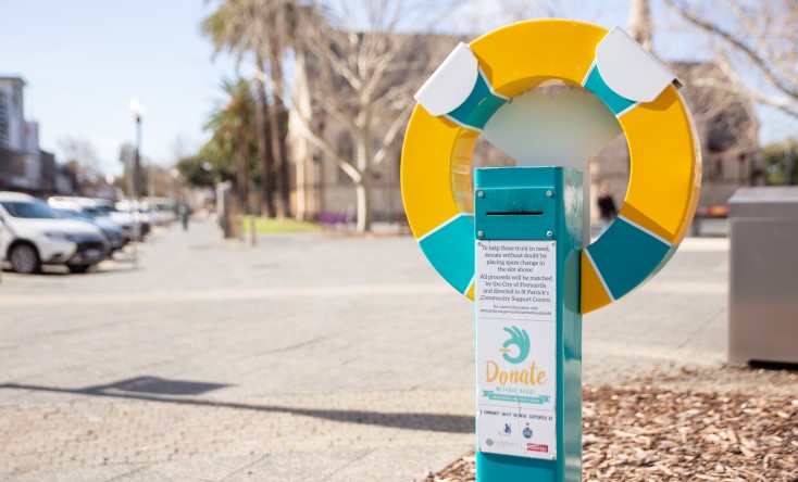 A yellow and green donation station shaped like a life preserver stands on a city sidewalk, with parked cars and trees visible in the background. A sign on the station encourages donations to support people in need.