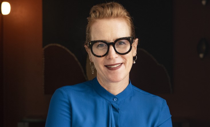 A woman with light skin and red hair, wearing black-rimmed glasses and a bright blue blouse, smiles at the camera. The background is dark with a lit wall sconce on the left.