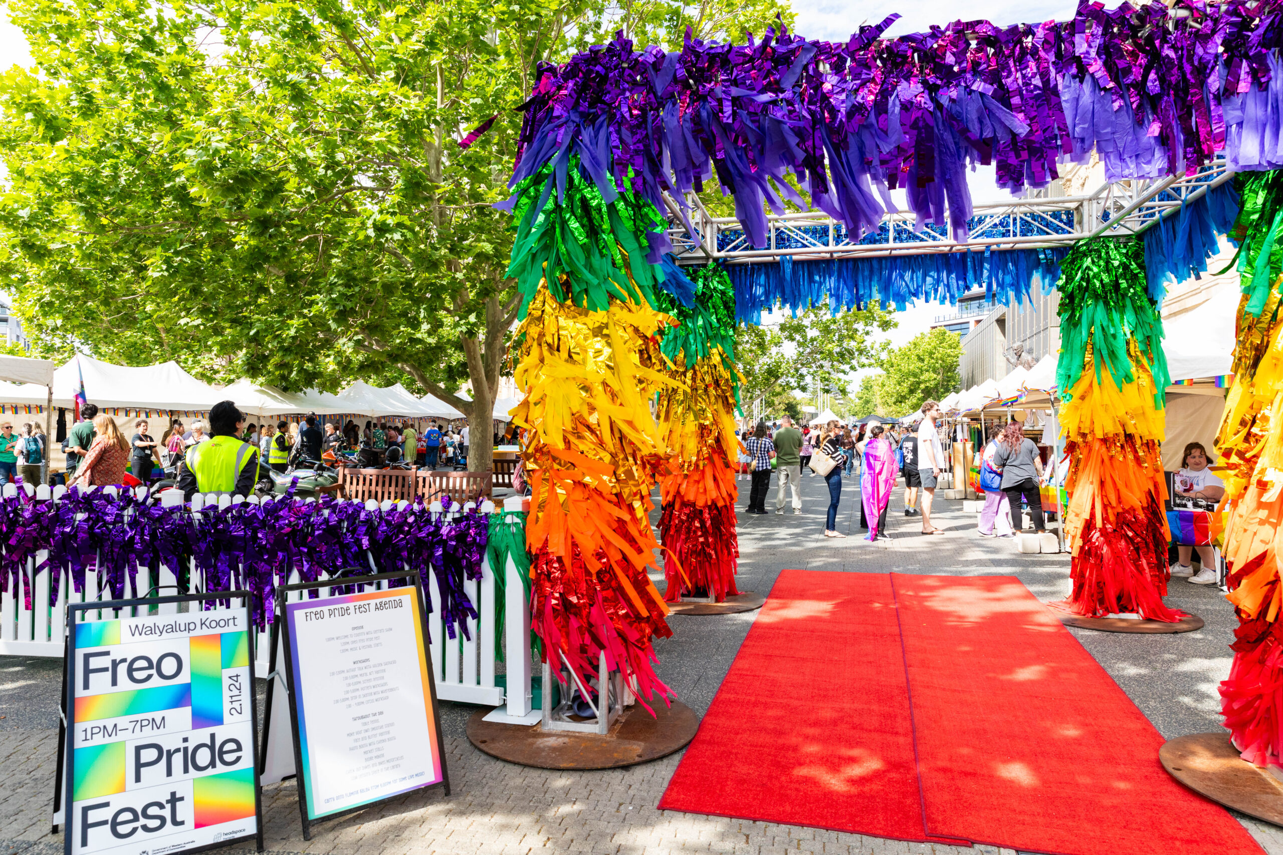 A vibrant outdoor pride festival entrance with rainbow-colored streamers, a red carpet, and a sign reading Freo Pride Fest. People gather under trees and white tents in the background.