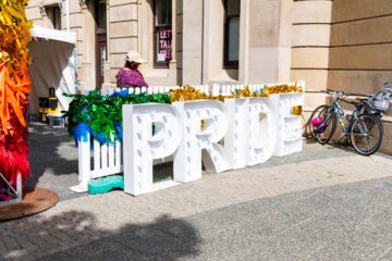 Large white letters spelling PRIDE are decorated with rainbow and gold tinsel, set up on a sidewalk in front of a stone building. A person in a hat stands nearby, and two bicycles are parked to the right.