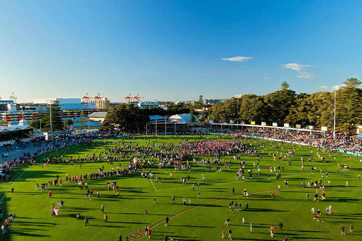 A large crowd of people spread across a green sports field under a clear blue sky, with trees, stadium seating, and buildings visible in the background.