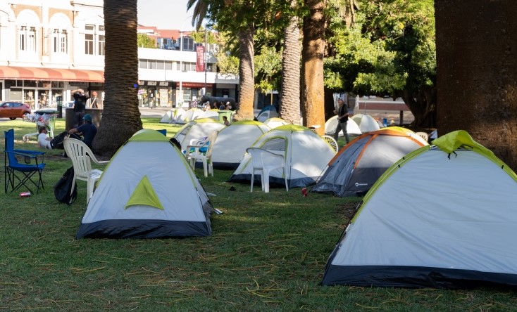 Several small tents are set up on a grassy area under palm trees, with people sitting nearby on white plastic chairs. Buildings are visible in the background.
