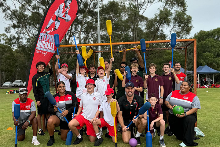 A diverse group of people, including kids and adults, pose together outdoors on a sports field holding foam cricket bats. They are smiling, with trees and a red Stephen Michael Foundation banner in the background.