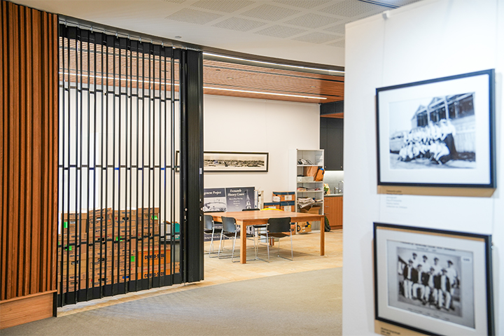 A modern interior featuring a gallery with framed black-and-white photos on the wall and an adjacent office area with a wooden table, chairs, and shelves, separated by vertical wooden slats.