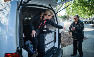 A woman sitting in the back of a van hands an item to another woman standing outside on a sidewalk. The vans back doors are open, revealing organized drawers and supplies inside. Trees and buildings are visible in the background.