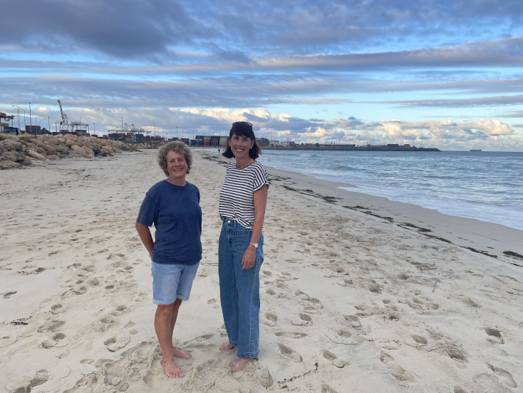 Two women stand barefoot on a sandy beach, smiling at the camera. The sky is partly cloudy with blue patches, and the ocean and distant buildings are visible in the background.