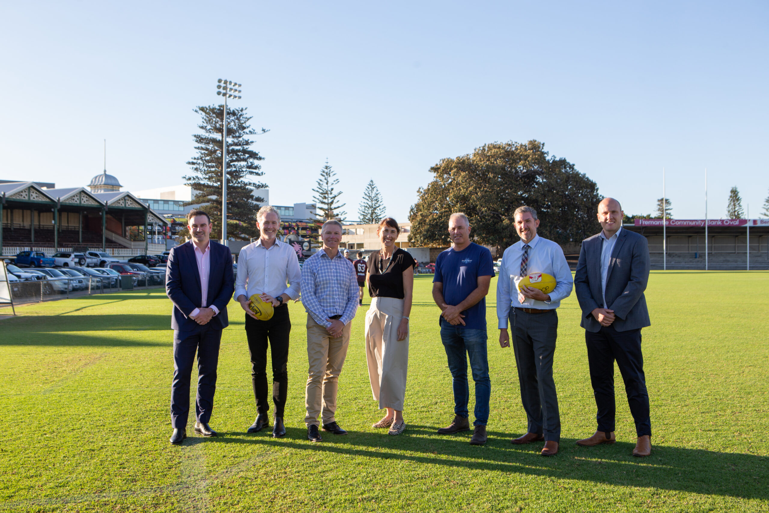 Seven people stand in a row on a grass sports field in daylight, with two holding yellow Australian footballs. Stadium seating, trees, buildings, and parked cars are visible in the background under a clear sky.