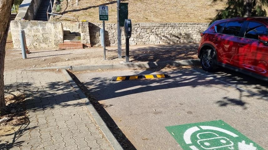 A parking space with an electric vehicle charging station, a yellow speed bump, and a red car parked nearby. The ground has a green EV charging symbol and a stone staircase is in the background.