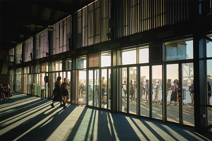 People stand in line outside large glass doors as sunlight streams into a spacious indoor area, casting long shadows across the floor. The scene appears to be at a terminal or a waiting area.