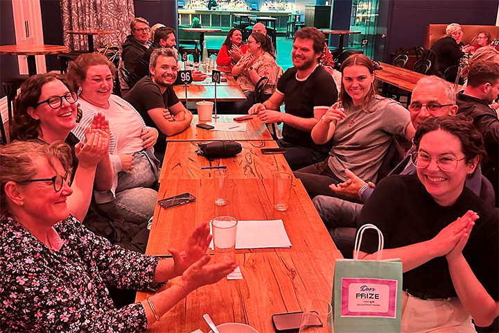 A group of people sit around a long table at a restaurant, smiling and clapping. A woman in the foreground holds a green prize bag labeled Prize. Plates, drinks, and papers are on the table. The atmosphere is cheerful.