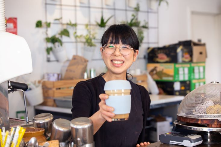 A smiling barista with glasses holds out a reusable coffee cup in a cozy cafe, standing behind the counter with coffee-making equipment and pastries visible.