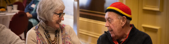 An older woman with gray hair and glasses smiles at an older man wearing a red and yellow knit hat, as they sit and chat together indoors.