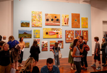 People gather in an art gallery, observing colorful abstract and landscape paintings displayed on a light blue wall. The room has wooden floors, and visitors are engaged in conversation and viewing the artwork.