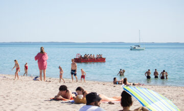 People relax on a sandy beach and swim in the sea. Some are sunbathing, others wade in the water. A group is riding a red boat, and a sailboat is anchored farther out on the calm, blue ocean.