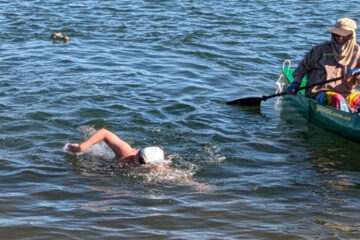 A swimmer wearing a white swim cap swims freestyle in open water next to a person in a green kayak on a sunny day.