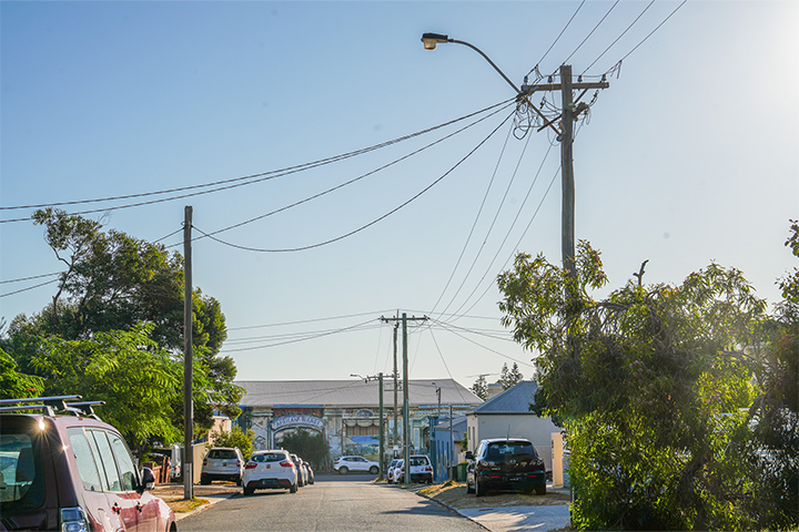 A suburban street with cars parked along the sides, utility poles and wires overhead, trees, and houses under a clear blue sky in daylight.