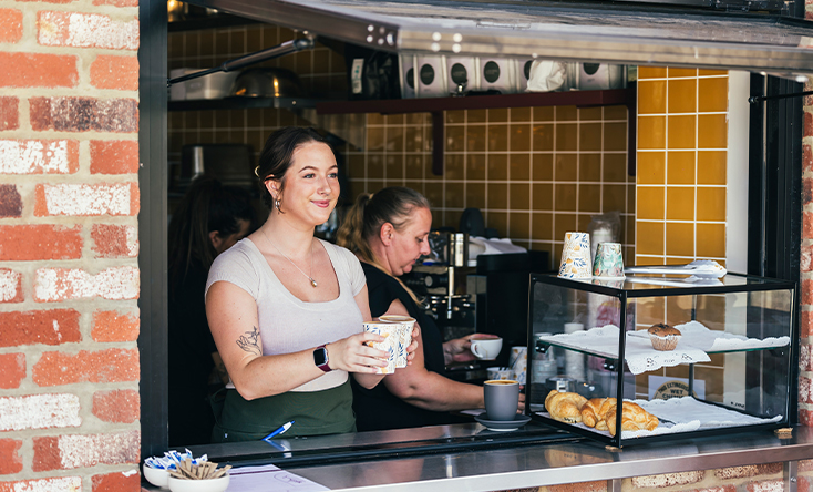 A smiling barista stands at a café window holding a takeaway coffee cup, with pastries displayed nearby and another woman working behind her. The café has yellow tiled walls and a brick exterior.