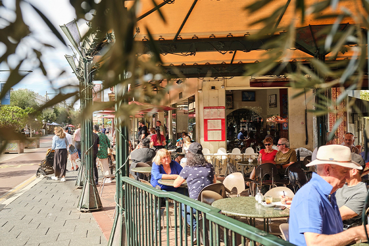 People sit at outdoor tables under a large canopy at a busy café on a sunny day. Some are eating or chatting, while others walk by on the sidewalk. Green railings and leafy plants frame the lively scene.