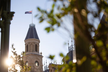 A historic clock tower with a flag on top is seen through leafy branches, with sunlight shining from the left side of the image. The sky is clear and blue.