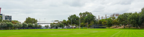A wide, green sports field with rugby goalposts, surrounded by trees and buildings under a cloudy sky. The area appears well-maintained and open.