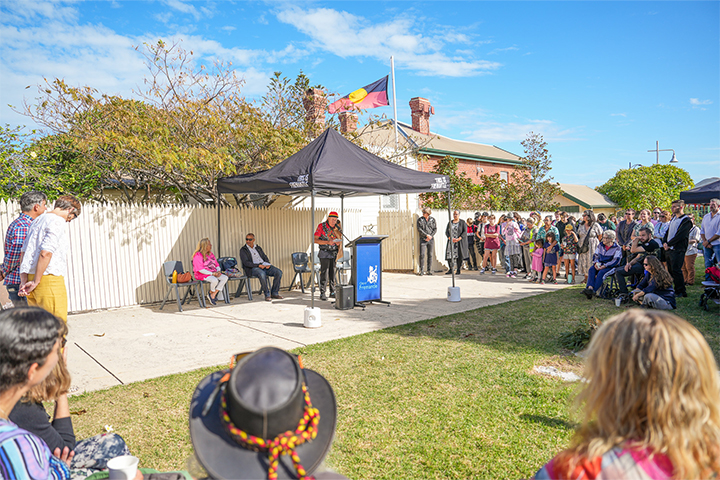 A crowd gathers outdoors around a speaker standing at a podium under a black tent. The Aboriginal flag flies on a pole above, and people are seated and standing, listening to the speech on a sunny day.