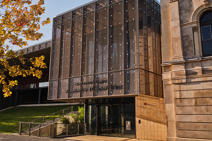 Modern building facade with perforated metal panels reading City of Fremantle Walyalup Civic Centre, next to an older stone structure; a tree with yellow leaves is on the left, and sunlight casts warm tones.